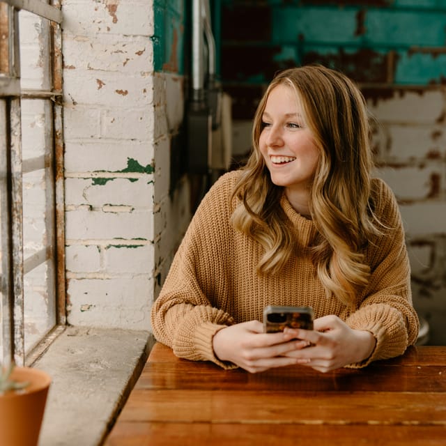 A young woman at a wooden table, smiling, with her phone in her hands.