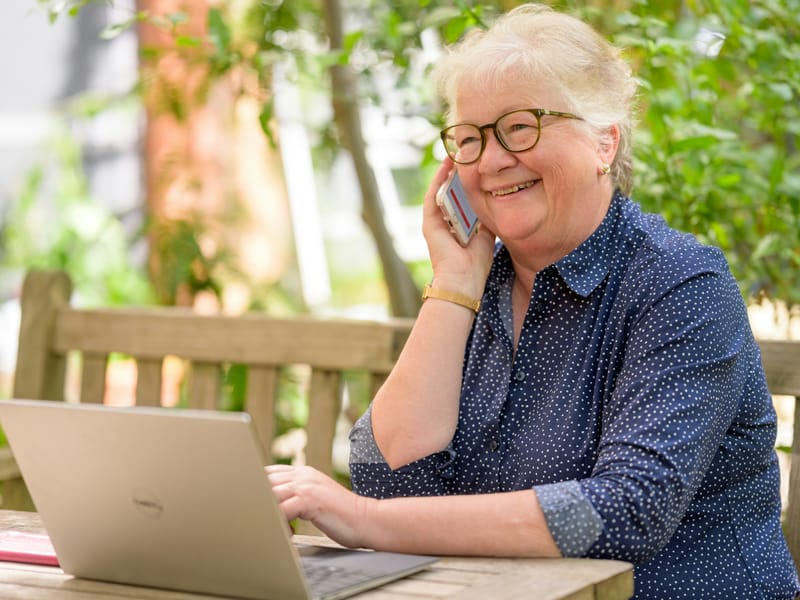 A woman at an outdoor table, on a phone call with a laptop open in front of her.