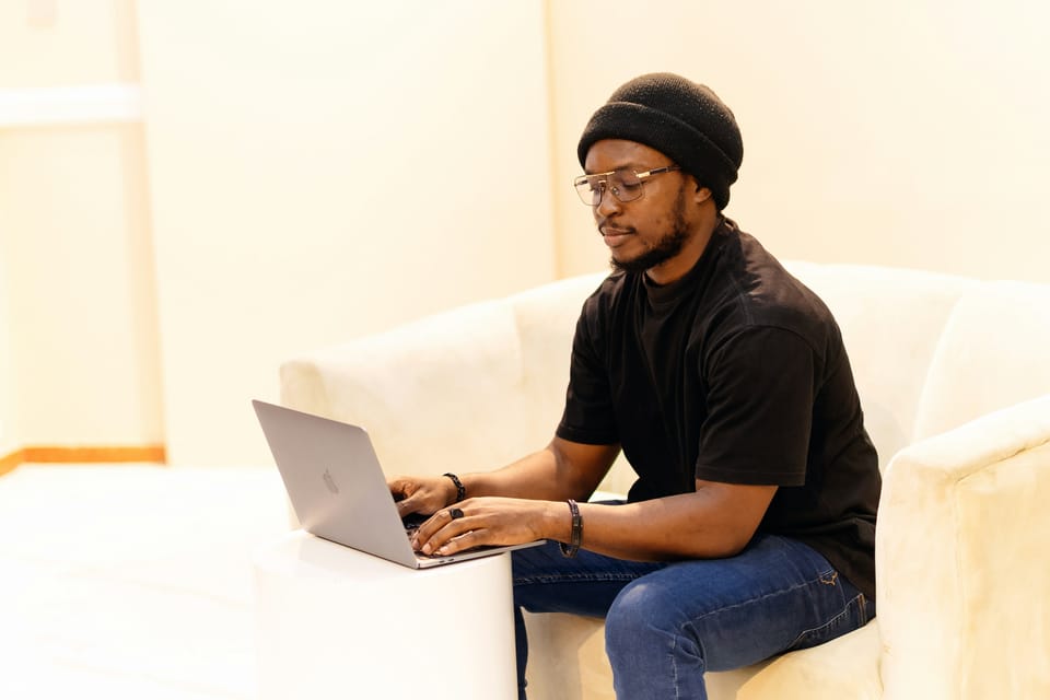 A man in a beanie on a sofa, working at his laptop.